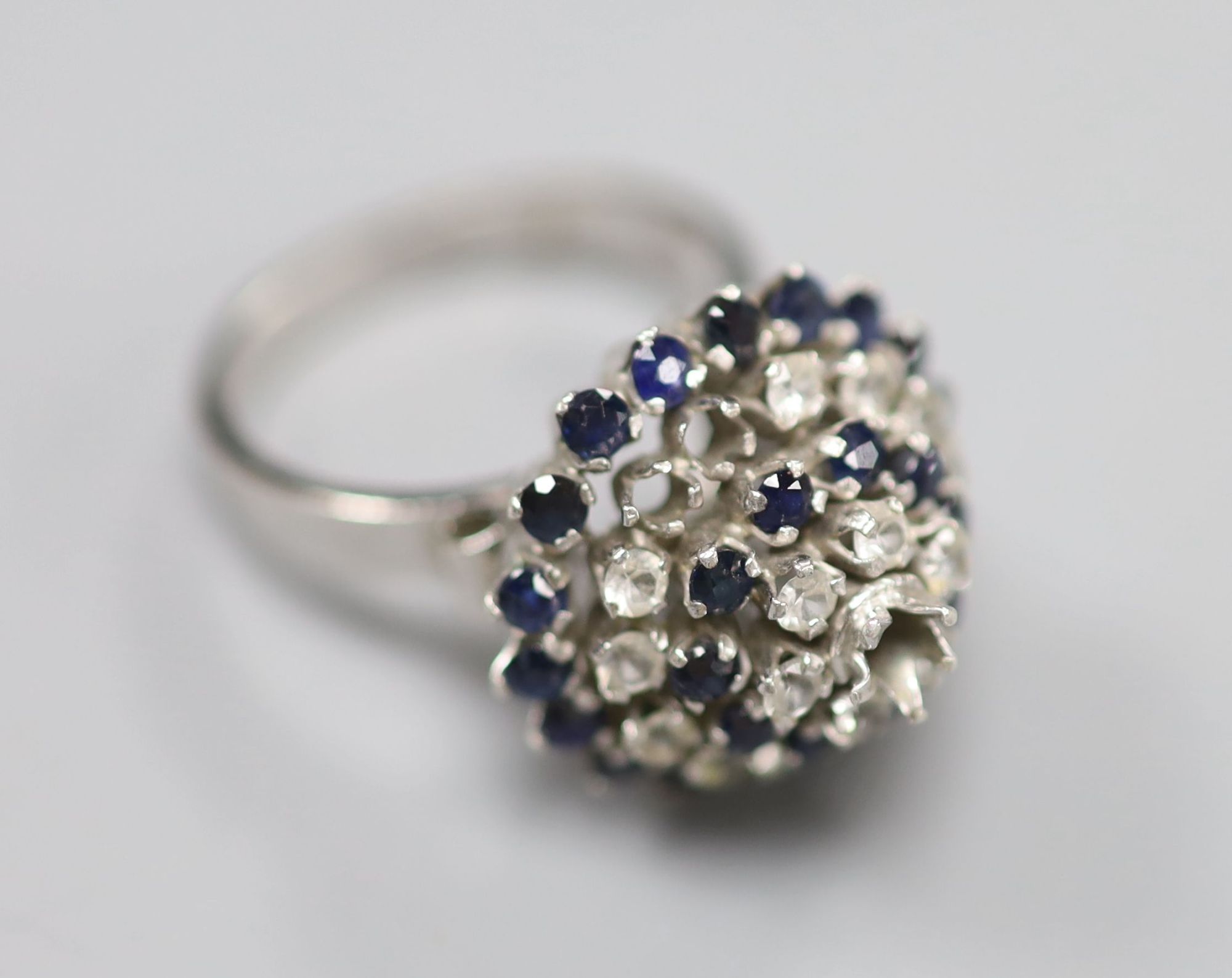 A white metal (stamped 18k) and two colour sapphire? set raised cluster ring, size K/L, gross 5.2 grams (one stone missing).